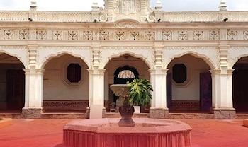 Un patio interior con un edificio de fachada blanca, arcos, columnas, una fuente de piedra con planta y pavimento rojizo