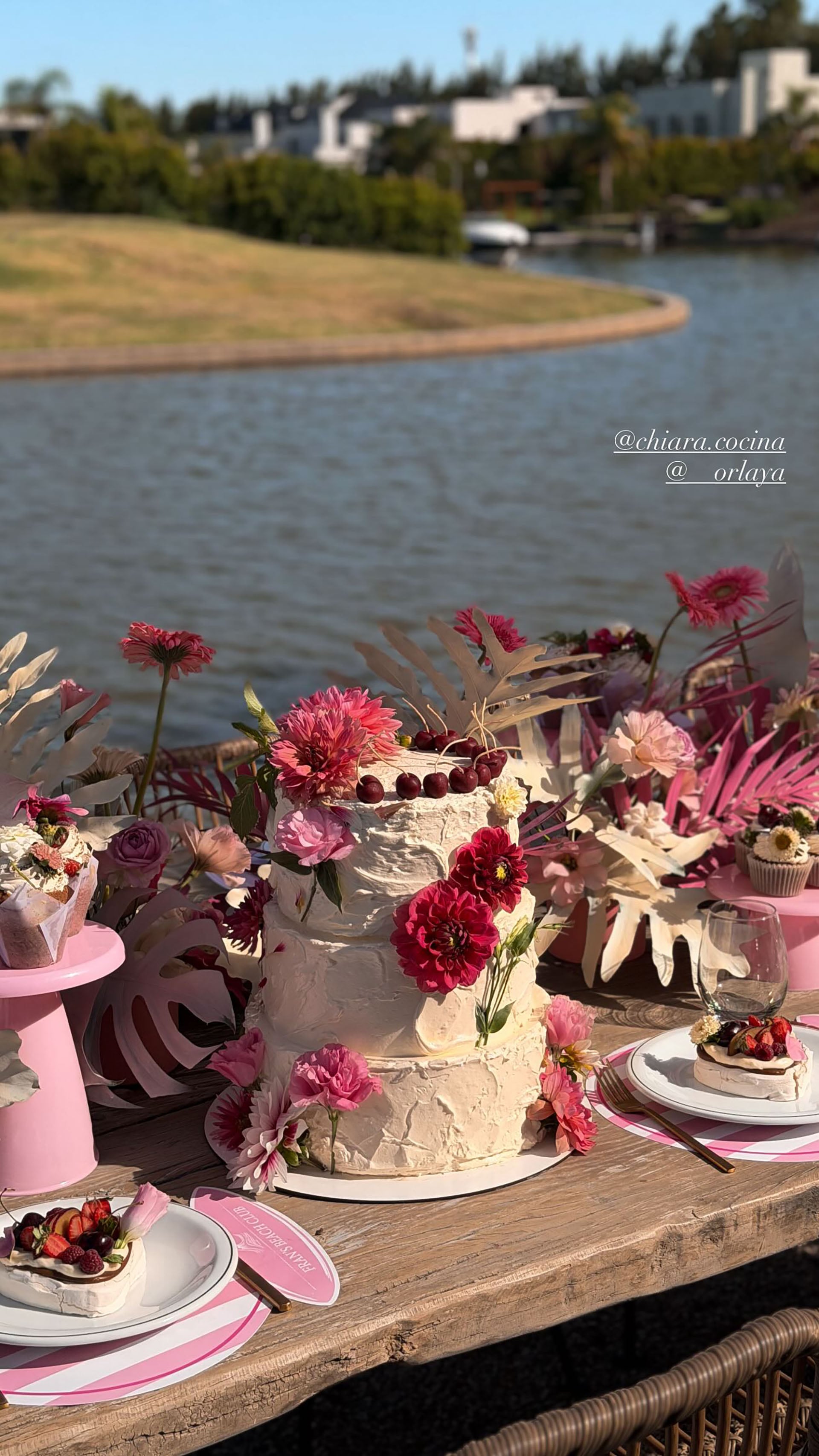 La torta de tres pisos decorada con flores frescas y 11 cerezas ocupa el centro de la mesa principal, rodeada de postres y vajilla en tonos pastel (Instagram)