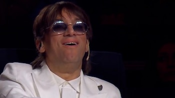Lady M, sonriendo, con cabello castaño y gafas de sol redondas y brillantes. Lleva chaqueta blanca, camisa blanca y audífono en la oreja. Fondo oscuro