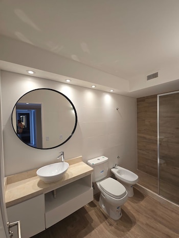 Vista de un baño moderno con lavabo ovalado sobre encimera de mármol, inodoro, bidé y cabina de ducha con pared de madera