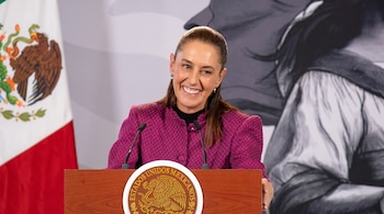 Claudia Sheinbaum sonríe detrás de un podio con el escudo de México, con la bandera nacional al fondo.