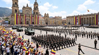 Plaza de Bolívar con desfile militar, soldados, vehículos blindados, multitud con banderas de Colombia, edificios decorados y confeti.