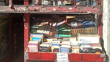 Vista frontal de un puesto callejero con estructura de metal rojo, lleno de libros, revistas y discos de vinilo apilados, con un cartel de precios visible