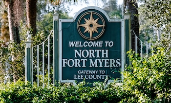 Un cartel verde con marco blanco, texto "Welcome to North Fort Myers Gateway to Lee County" y una rosa de los vientos dorada, rodeado de vegetación