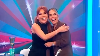 Dos mujeres sonríen ampliamente mientras se abrazan en un set de televisión con un fondo de colores vibrantes y luces brillantes. Hay un micrófono y una copa.