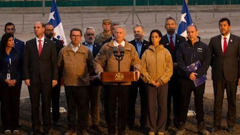 Un grupo de hombres, incluyendo militares y funcionarios, se congrega al aire libre en un paisaje desértico, bajo una bandera chilena. Miran un caballete