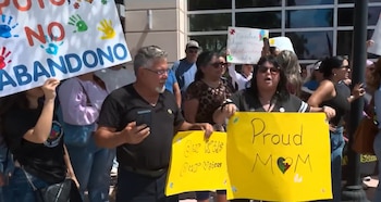 Um grupo de adultos protestando ao ar livre. Uma mulher segurando uma placa amarela dizendo