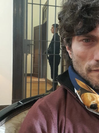 Primer plano del rostro de Fabio Di Tomaso con cabello rizado y barba, vistiendo una chaqueta granate. Al fondo, una figura en uniforme detrás de rejas metálicas
