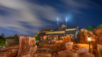 Edificio de Big Thunder Mountain Railroad iluminado de noche, con rocas y chimeneas emitiendo luz; el cielo nocturno con nubes alargadas