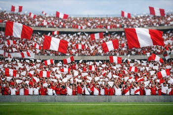 Vista horizontal de tribunas repletas de personas vestidas de blanco y rojo, ondeando numerosas banderas peruanas grandes y pequeñas y pañuelos blancos.