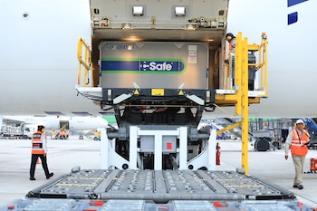 Avión de carga blanco, contenedor de carga gris con el logo "Safe", rampa de carga, operarios de aeropuerto con chalecos de seguridad, vehículos de apoyo en pista