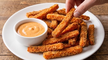 Mano sostiene bastón empanizado, plato blanco con bastones empanizados, cuenco blanco con salsa naranja, superficie de madera.
