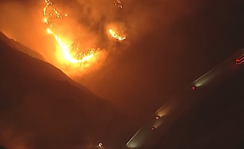 Vista aérea nocturna de un incendio forestal masivo en una ladera, con llamas brillantes y humo denso, y una carretera oscura con vehículos