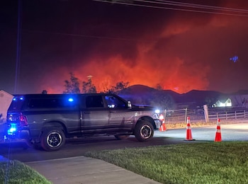 Camioneta negra con luces azules intermitentes en la calle. Detrás se observa un gran incendio rojo, árboles oscuros y montañas, bajo un cielo nocturno