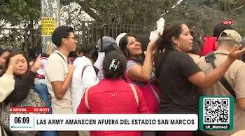 Varias personas agrupadas en una fila sobre la acera frente a una reja metálica con un rótulo informativo de televisión