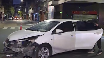 Automóvil blanco con el frente destrozado y capó levantado, detenido en una calle oscura. Una puerta trasera está abierta, cono de tráfico rojo cercano