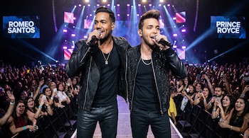 Romeo Santos y Prince Royce cantando con micrófonos en un escenario iluminado por luces moradas y azules. Visten de negro frente a una multitud de fans.