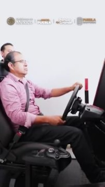 Hombre sentado en un simulador de manejo. Viste camisa rosa y cinturón de seguridad. Se ven logotipos del Gobierno de México y Puebla en la parte superior