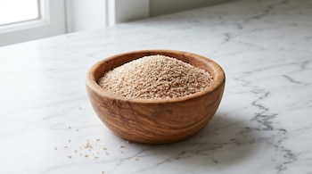 Un cuenco de madera claro lleno de pequeñas semillas de psyllium beige sobre una mesada de mármol blanco y gris, con algunas semillas esparcidas.