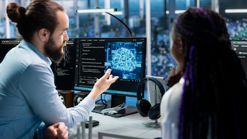 Un hombre gesticula frente a un monitor con el dibujo digital de un cerebro y las letras AI mientras una mujer escucha a su lado