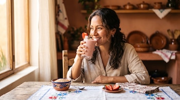 Una mujer sostiene un vaso con batido rosado junto a un tazón de barro con granos, fresas cortadas y un mantel bordado.