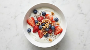 Vista aérea de un tazón blanco con yogur natural, fresas, arándanos, semillas de chía y nueces. Se apoya sobre una superficie de mármol claro.