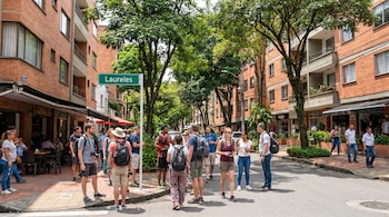 Vista de una calle urbana con edificios de ladrillo, árboles frondosos y un grupo de personas, muchos de ellos turistas, de pie cerca de un letrero que dice "Laureles".