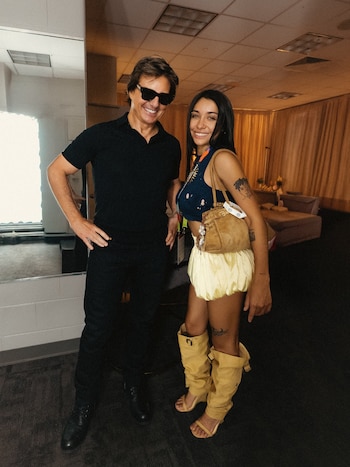 Tom Cruise con gafas de sol, polo negro y pantalón, y María Becerra con top azul, minifalda amarilla, botas y bolso, posan en un interior