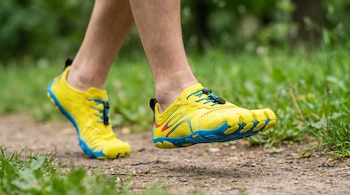 Primer plano de dos pies humanos calzados con zapatillas amarillas y azules de dedos separados caminando en un sendero de tierra. Fondo verde borroso.