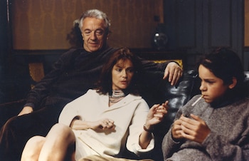 Tres personas sentadas en un sofá oscuro: un hombre mayor de cabello gris detrás de una mujer de cabello castaño y un joven con cigarrillo