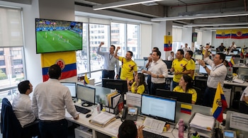 Una oficina con trabajadores viendo un partido de fútbol en una pantalla grande; hay banderas de Colombia, computadoras y escritorios con papeles.
