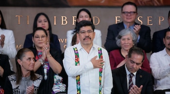 Un hombre con camisa blanca bordada permanece de pie con la mano sobre el pecho ante varias personas sentadas que aplauden.