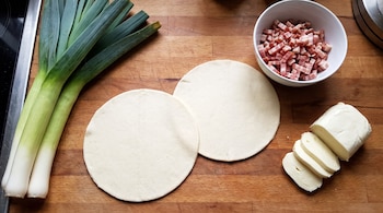 Tres puerros, dos discos de masa cruda, un cuenco con panceta en cubos y un bloque de mozzarella cortada en rodajas sobre una tabla de madera.