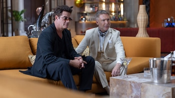 Dos hombres sentados en un sofá naranja. El de la izquierda viste traje oscuro y gafas de sol, el de la derecha traje claro. Un cóctel y un cubo de hielo están en la mesa