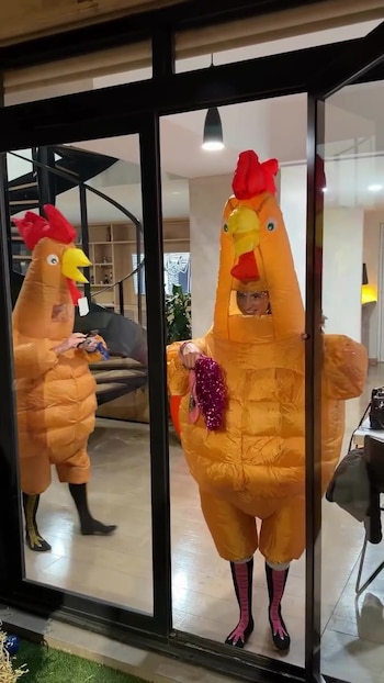 Dos personas con disfraces de pollo inflables naranjas. Una sostiene un objeto brillante rosado. Reflejo en puerta de vidrio. Escalera de caracol visible