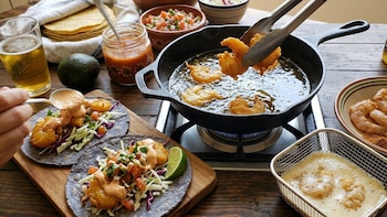 Primer plano de manos friendo camarones capeados en una sartén de hierro y sirviendo tacos con lechuga, pico de gallo y salsa. Tortillas, aguacate y cerveza en la mesa