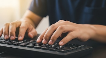 Primer plano de dos manos con piel clara escribiendo sobre un teclado de computadora negro. La persona viste una camisa azul oscuro. El fondo está iluminado con luz suave.
