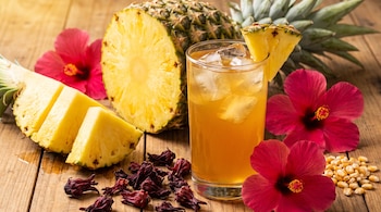 Vaso de tepache con hielo y rodaja de piña, piña a la mitad, trozos de piña, flores de hibisco rojas, flores de jamaica secas y granos de maíz.