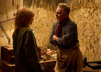 Hombre de camisa gris y pañuelo rojo con delantal beige dialoga con mujer de cabello rubio y chaqueta verde en estudio de arte con pinturas y lámpara