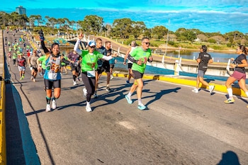 Grupo de personas corriendo en una carretera, con un puente sobre agua, árboles y un edificio al fondo bajo un cielo azul con nubes