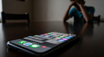 Primer plano de teléfono inteligente sobre una mesa oscura con notificaciones en pantalla; al fondo, silueta desenfocada de adolescente con la cabeza entre las manos.