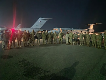 Grupo de militares con uniformes camuflados, uno con perro, de pie frente a dos aviones jet en la pista de un aeropuerto, de noche