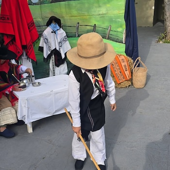 Niño vestido con sombrero de ala ancha, camisa blanca, chaleco negro y pantalón blanco, sosteniendo un palo de madera. Fondo con escenografía rural y otras personas