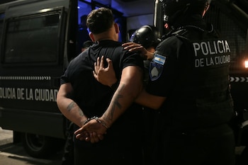 Un hombre esposado con las manos a la espalda es sujetado por un oficial de la Policía de la Ciudad ante un vehículo policial