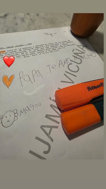 Primer plano de una hoja de papel con texto impreso, la frase manuscrita "Papa Te Amo" y un dibujo infantil, junto a dos marcadores naranjas