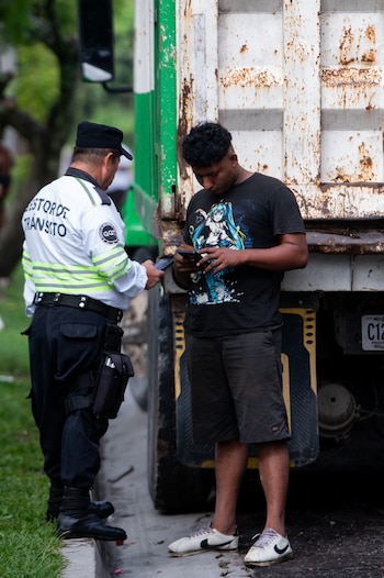 Un gestor de tránsito parado junto a un joven con camiseta negra que consulta su teléfono móvil cerca de un camión