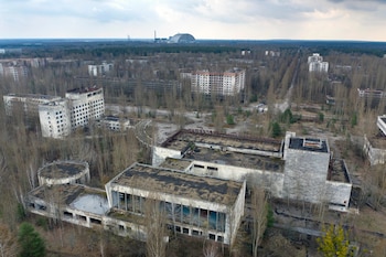Vista aérea de una ciudad desolada con edificios deteriorados y extensos árboles sin hojas. Al fondo, una gran estructura abovedada de acero gris bajo cielo nublado