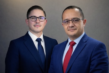 Dos hombres con trajes azul oscuro y gafas posan de frente. El de la izquierda lleva corbata azul, el de la derecha corbata roja, ambos con fondo gris.