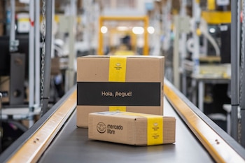 Dos paquetes de cartón de Mercado Libre, con cinta amarilla y el logo, uno grande y otro pequeño, se mueven sobre una cinta transportadora gris en un almacén. El paquete grande dice "Hola, ¡llegué!"