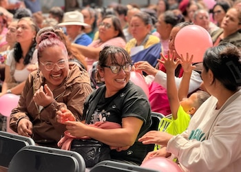 Varias mujeres sentadas en sillas sonríen y conversan rodeadas de globos rosados entre una multitud